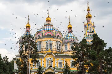 View of Panfilov Park in Almaty showcasing the iconic Zenkov Cathedral surrounded by lush greenery, vibrant red flower beds, and a tree-lined pathway, capturing the park's serene beauty and cultural significance.