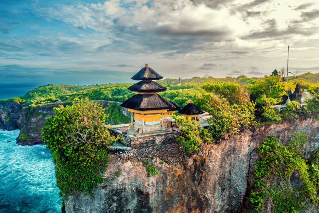Uluwatu Temple perched on a dramatic cliff overlooking the Indian Ocean, surrounded by lush greenery under a partly cloudy sky in Bali.